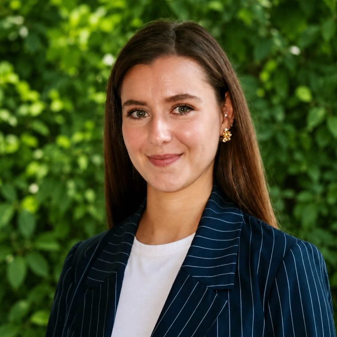 Portrait of a smiling woman with long brown hair wearing a navy pinstripe blazer outdoors among green foliage.