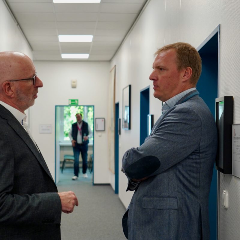Two men in business attire talking in a bright office hallway; one holds a jacket while the other gestures.