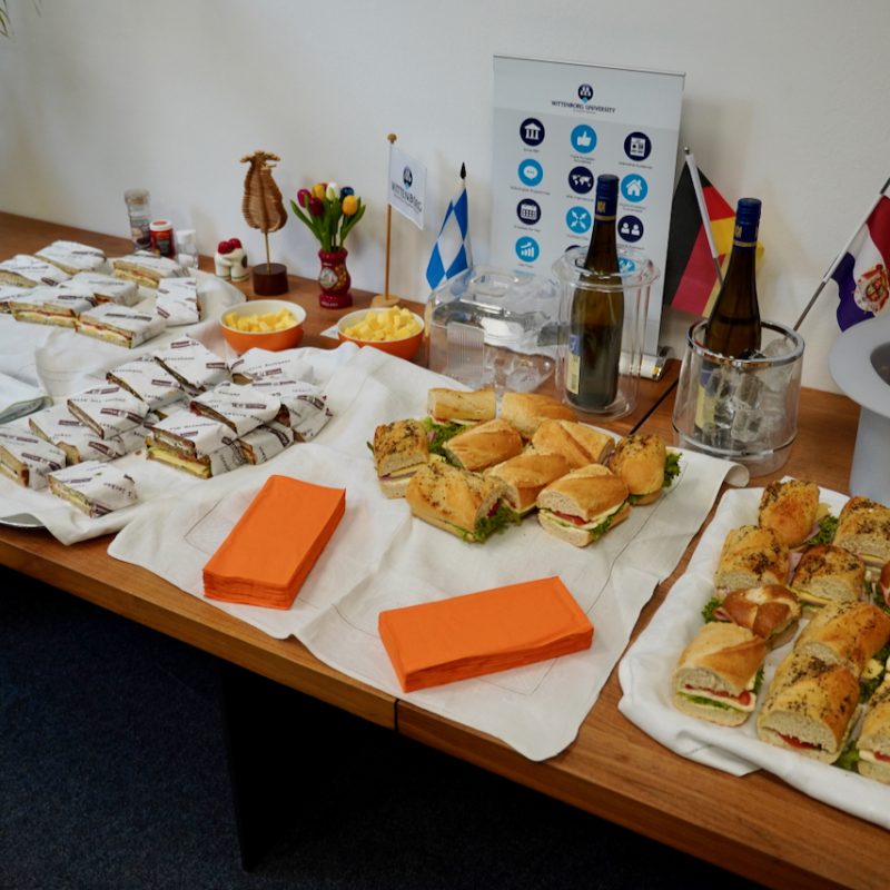 Buffet table with assorted finger sandwiches, wraps, cheese cubes, and orange napkins at a corporate event setup.