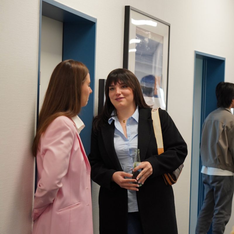 Two women chatting in an office hallway by a blue door; one wears a pink blazer, the other a black coat holding a bottle.