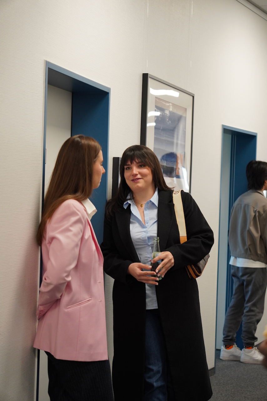 DHV00880 Two women chatting in an office hallway by a blue door; one wears a pink blazer, the other a black coat holding a bottle.