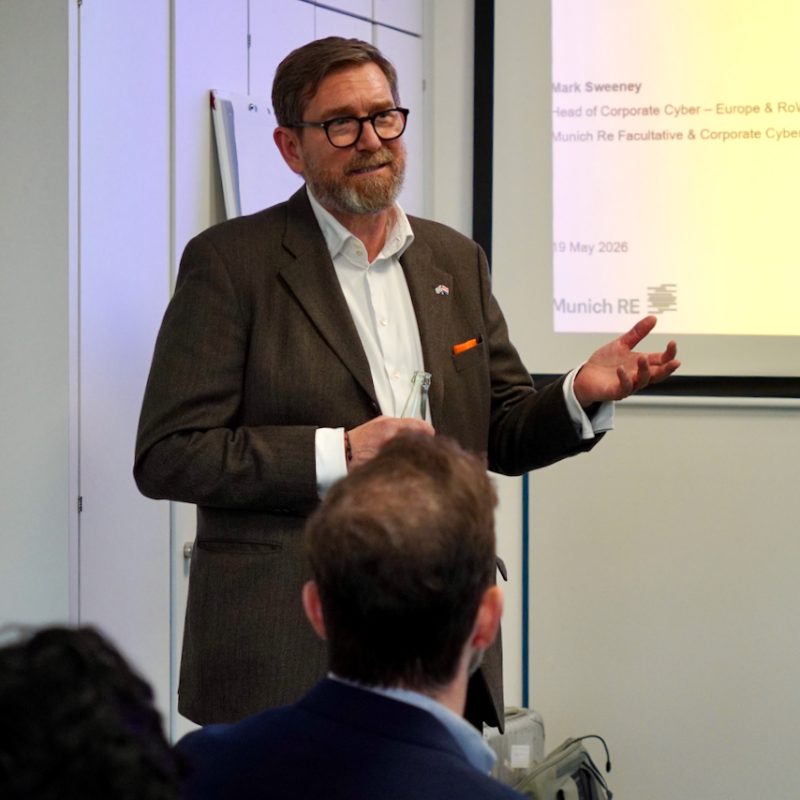 Speaker in a brown blazer and glasses presenting at a conference, gesturing toward a projected slide mentioning AI and cyber insurance.
