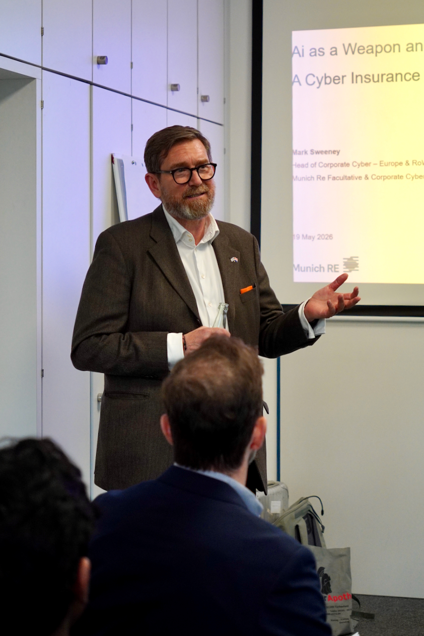 DHV00903 Speaker in a brown blazer and glasses presenting at a conference, gesturing toward a projected slide mentioning AI and cyber insurance.