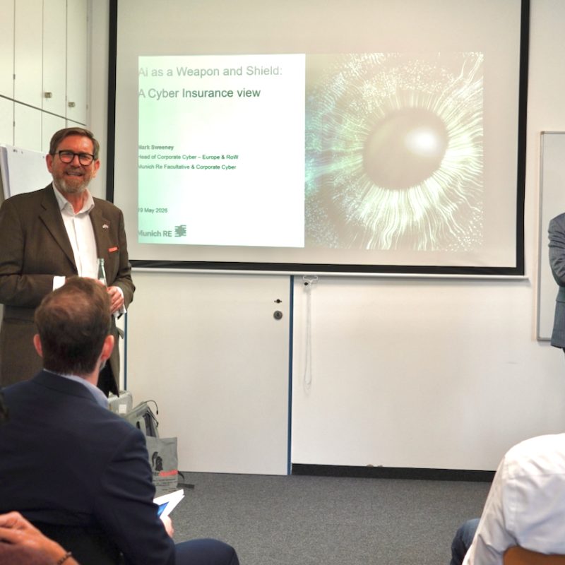 Two male presenters in a meeting room; one speaks at a projector screen while audience watches.