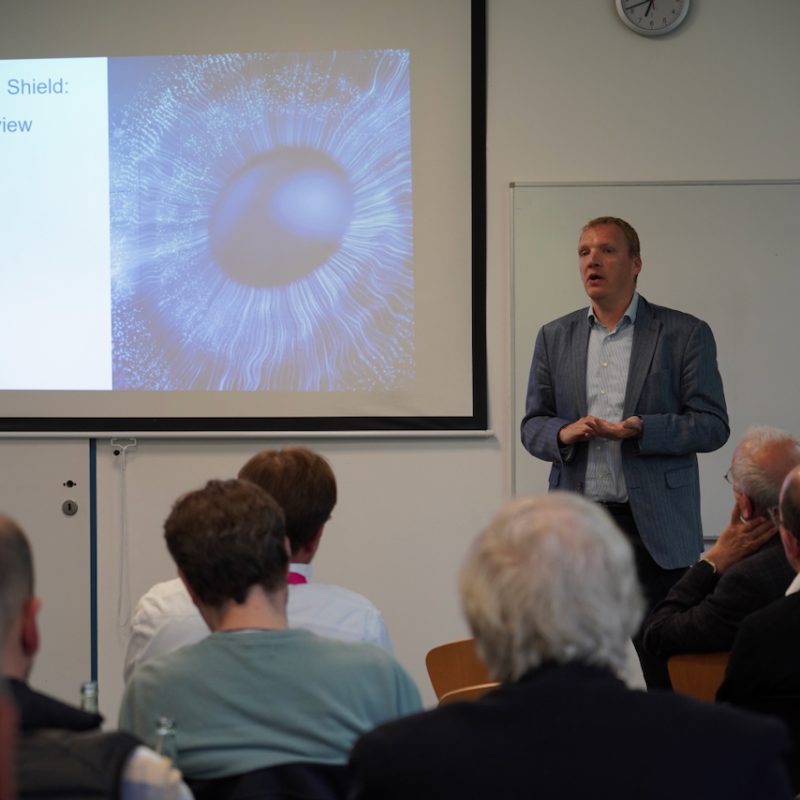 Male presenter in a blue blazer speaks to an audience as a slide shows 'AI as a Weapon and Shield: A Cyber Insurance view'