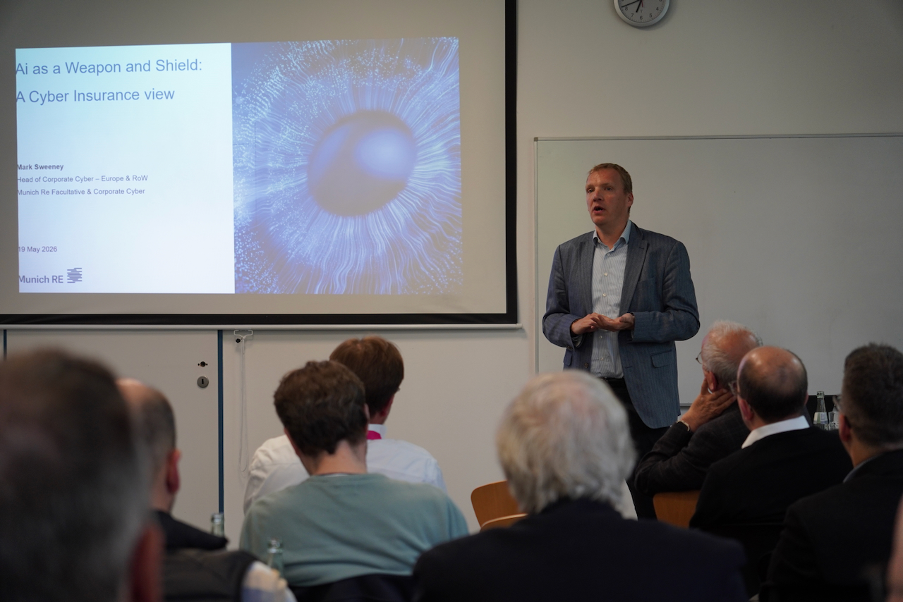 DHV00913 Male presenter in a blue blazer speaks to an audience as a slide shows 'AI as a Weapon and Shield: A Cyber Insurance view'