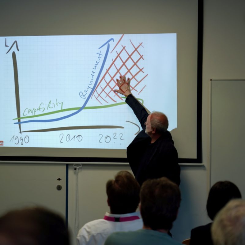 Older man lecturing, pointing at a projected graph on a screen showing a rising blue curve, a green capacity line, and a red hatch-filled area around 2020s.