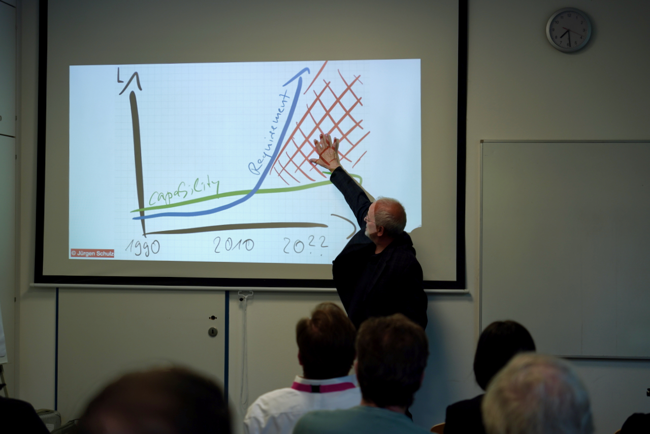 DHV00927 Older man lecturing, pointing at a projected graph on a screen showing a rising blue curve, a green capacity line, and a red hatch-filled area around 2020s.