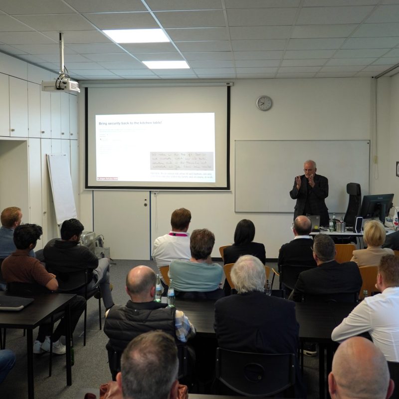 Audience seated in a classroom-style conference room, listening to a speaker at the front with a projected presentation on a screen and a whiteboard nearby.