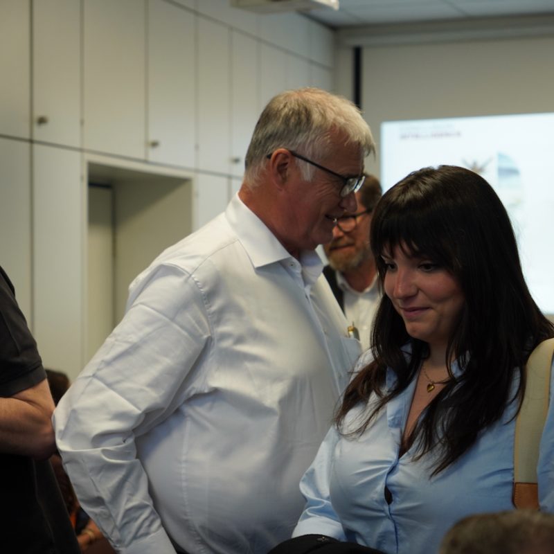 Older man in a white shirt smiles while talking to a woman in a blue blouse in a conference room, with a presentation screen in the background.