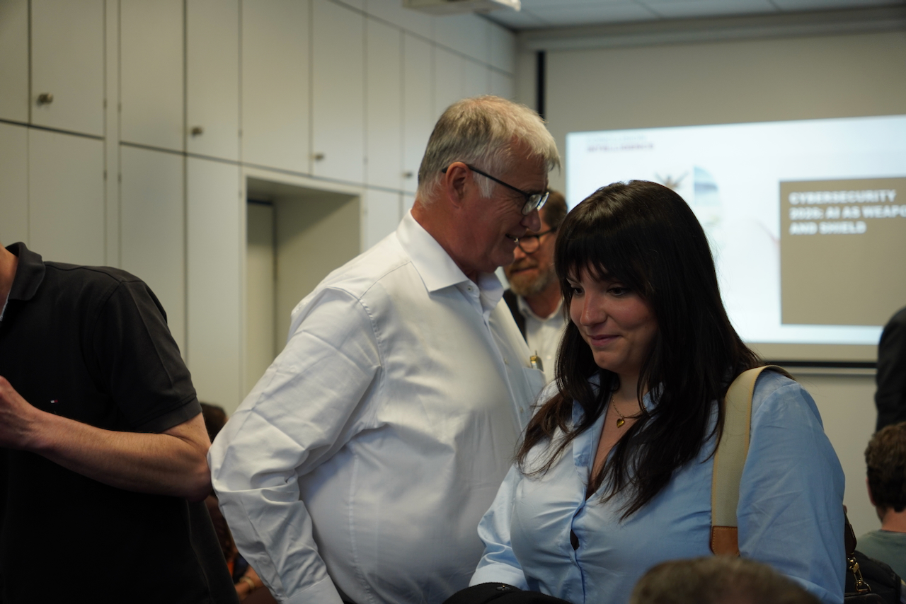 DHV00934 Older man in a white shirt smiles while talking to a woman in a blue blouse in a conference room, with a presentation screen in the background.
