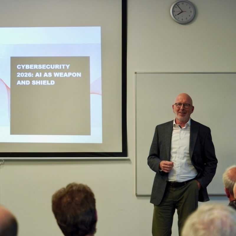 Bald man in a blazer presents a cybersecurity lecture beside a slide reading 'CYBERSECURITY 2026: AI AS WEAPON AND SHIELD'.