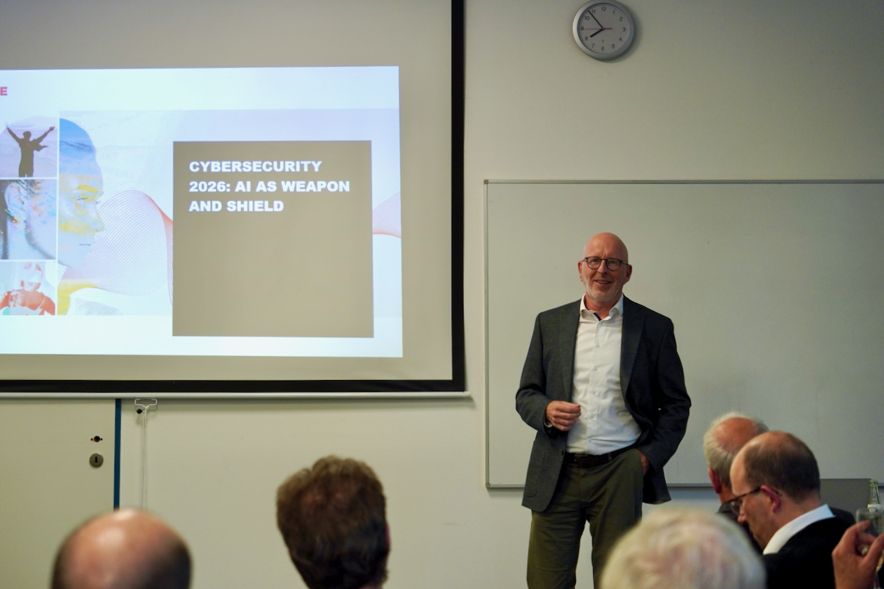 DHV00938 Bald man in a blazer presents a cybersecurity lecture beside a slide reading 'CYBERSECURITY 2026: AI AS WEAPON AND SHIELD'.