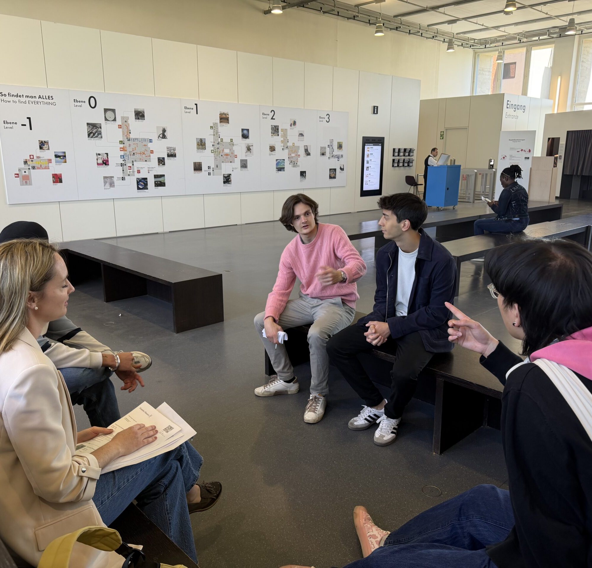 People seated on benches in a bright museum-like space, engaging in conversation with a few looking at each other and notes. A large wall with maps or posters is in the background.