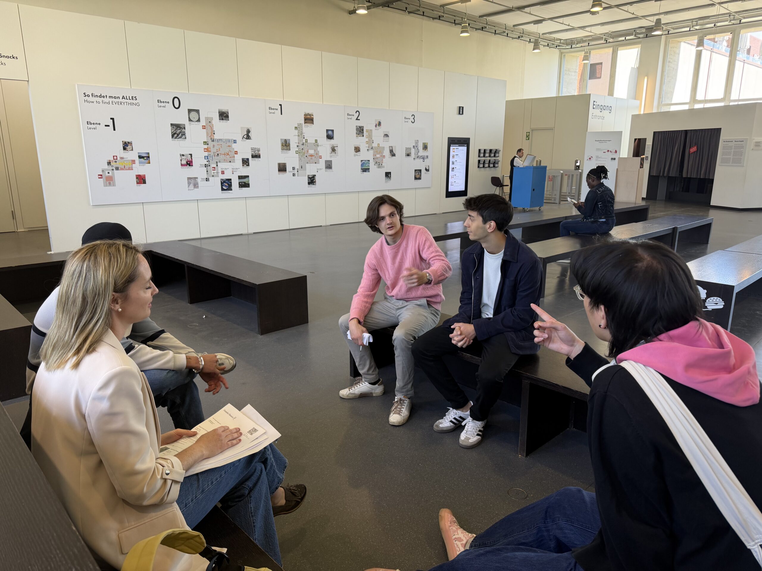 People seated on benches in a bright museum-like space, engaging in conversation with a few looking at each other and notes. A large wall with maps or posters is in the background.