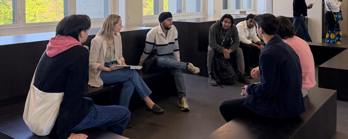 Group of diverse people sitting on black tiered benches in a bright, modern room, chatting together near large windows.