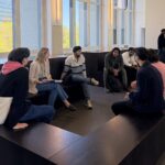 Group of diverse people sitting on black tiered benches in a bright, modern room, chatting together near large windows.