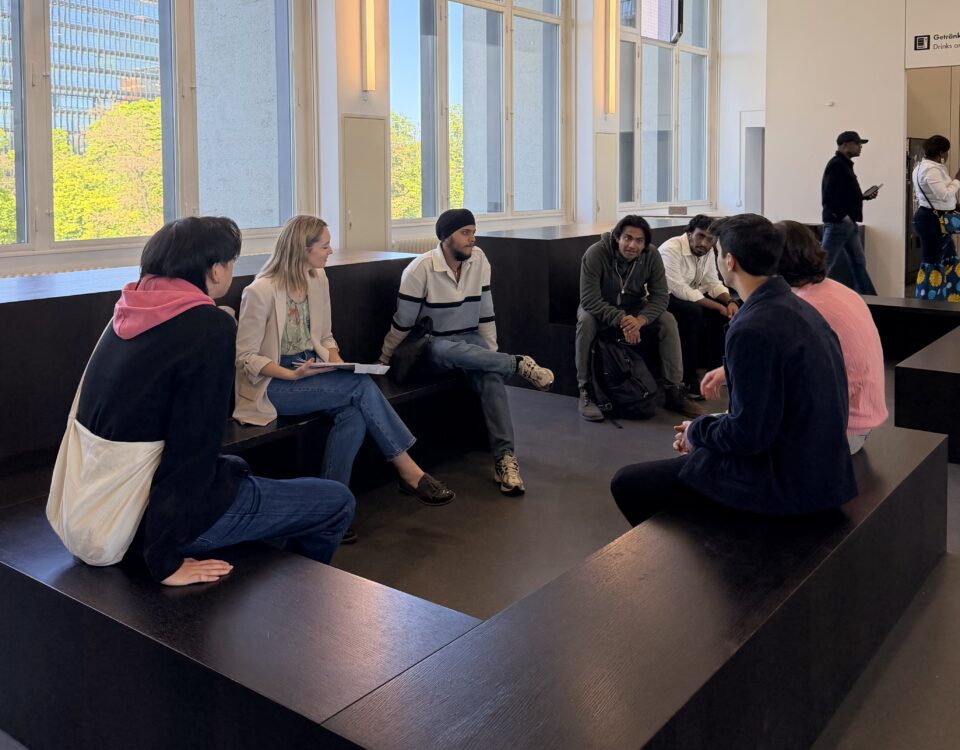 Group of diverse people sitting on black tiered benches in a bright, modern room, chatting together near large windows.