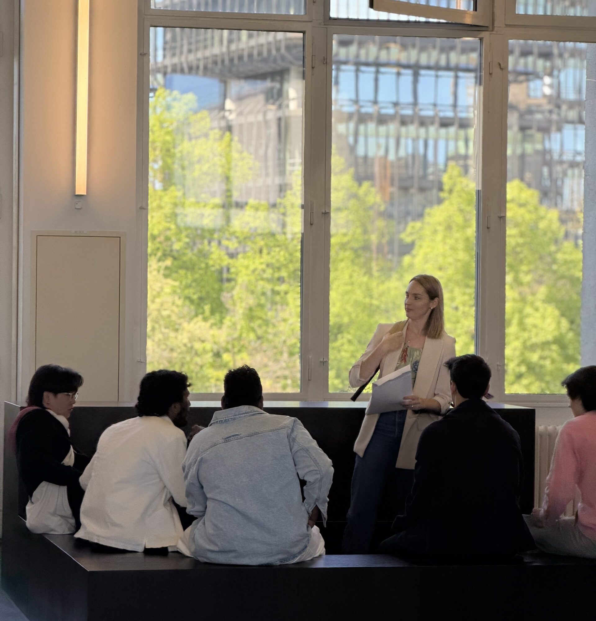 Woman standing with papers, presenting to a small audience in a bright room with large windows and a city view outside.