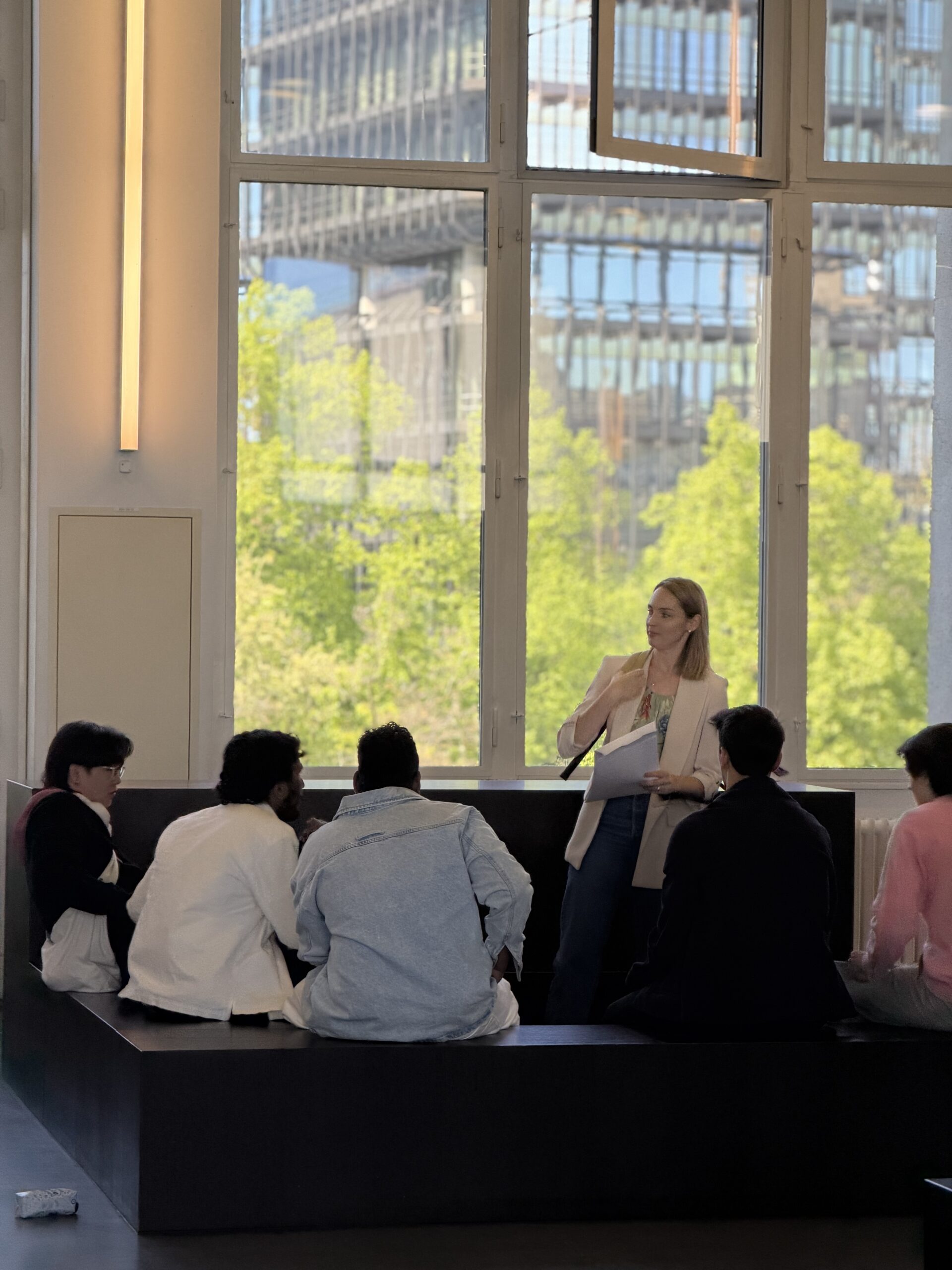 Woman standing with papers, presenting to a small audience in a bright room with large windows and a city view outside.