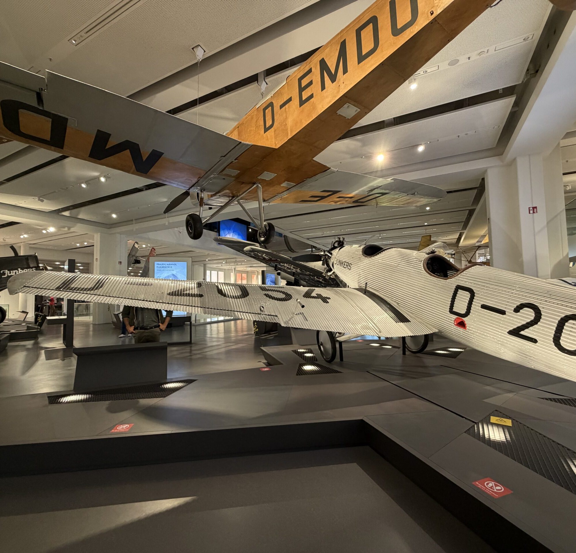 Indoor aviation museum showing vintage aircraft suspended from the ceiling, including a large wooden-winged plane marked 'D-EMD' above the viewer's head.