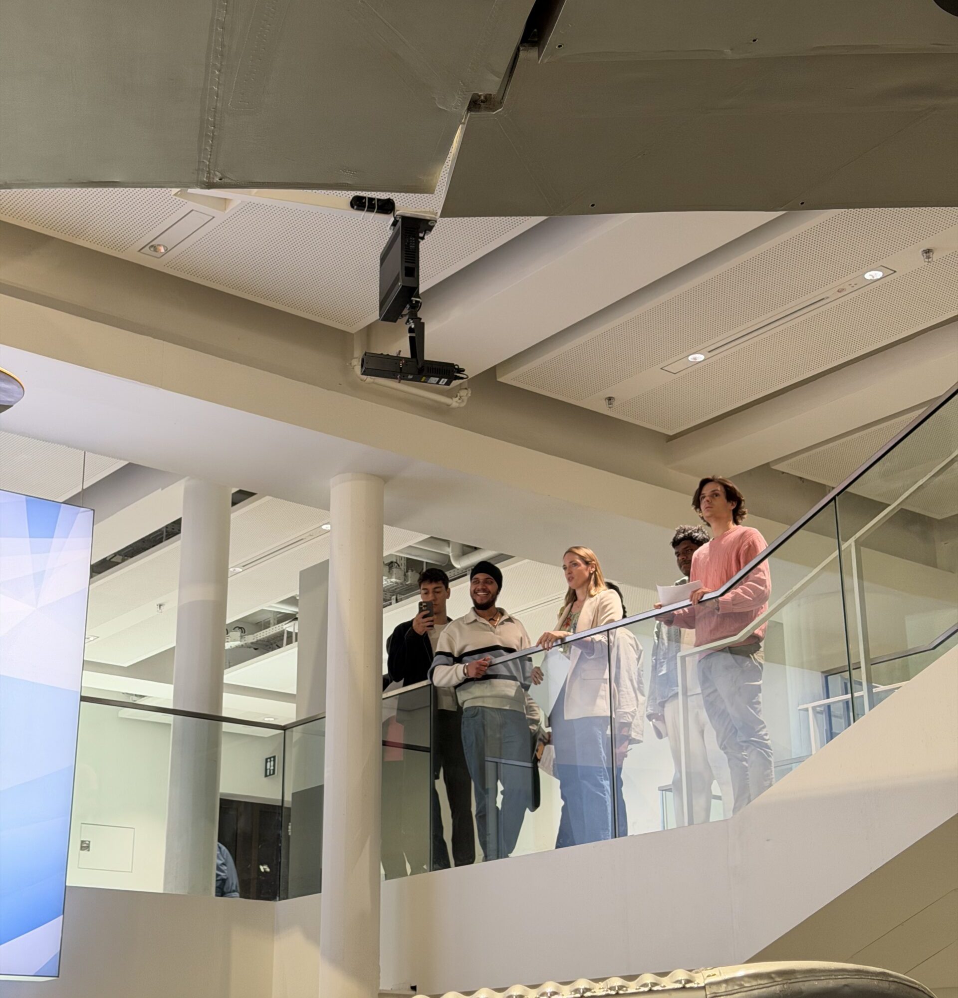 Group of people on a balcony looking at a large riveted metal aircraft fuselage hanging from the ceiling in a museum.