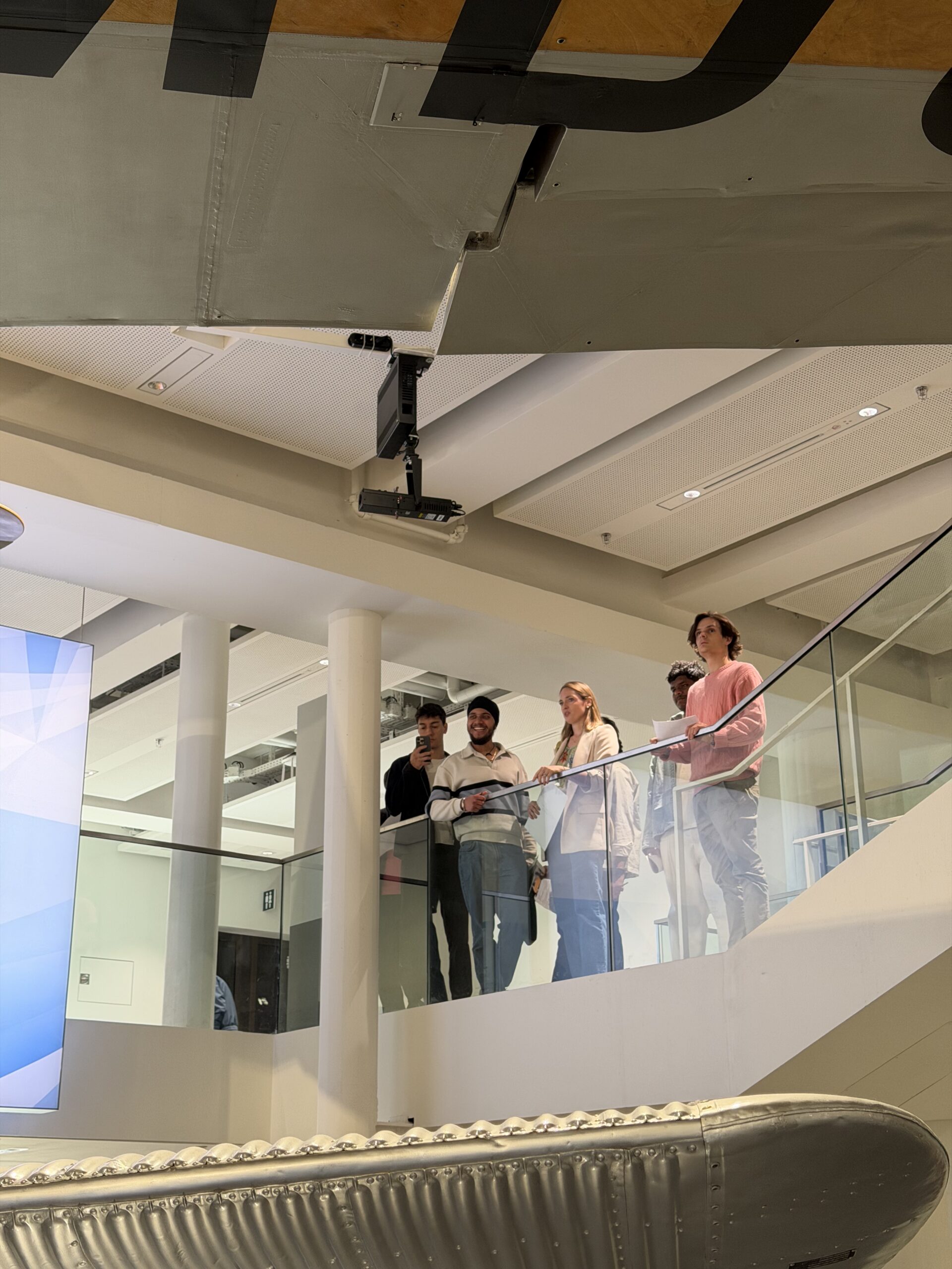 Group of people on a balcony looking at a large riveted metal aircraft fuselage hanging from the ceiling in a museum.