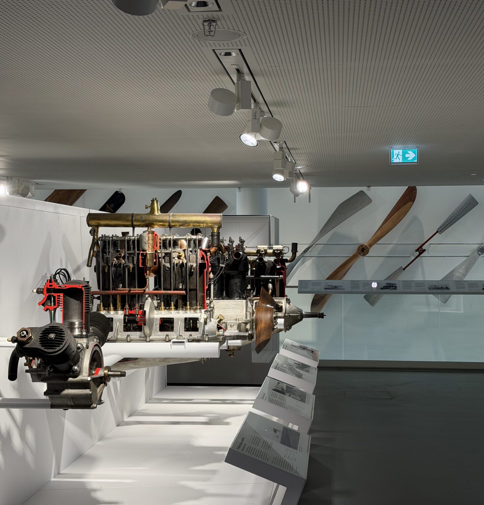 Suspended vintage aircraft engine on display with large wooden propeller blades mounted along a wall in a museum exhibit.