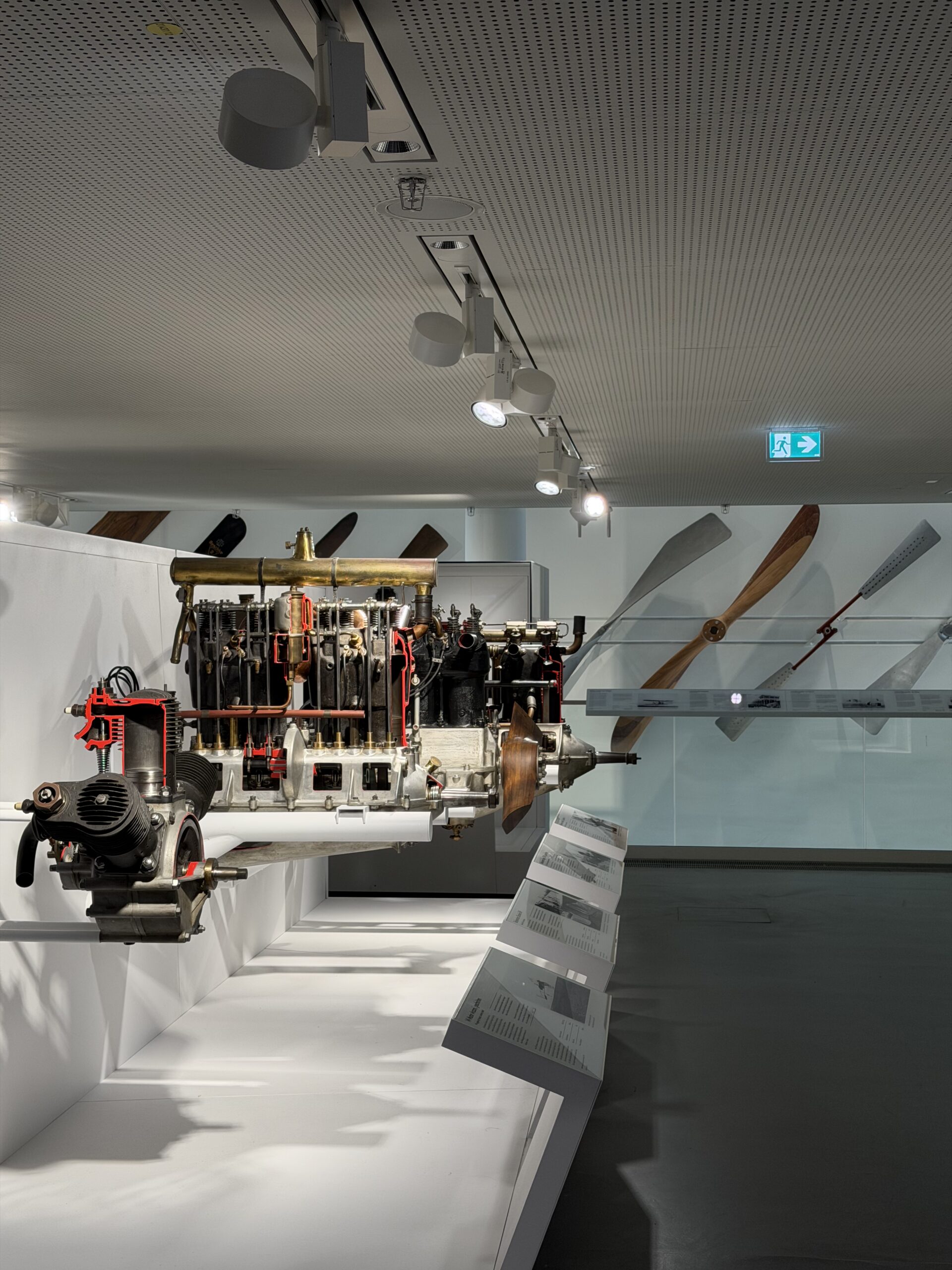 Suspended vintage aircraft engine on display with large wooden propeller blades mounted along a wall in a museum exhibit.