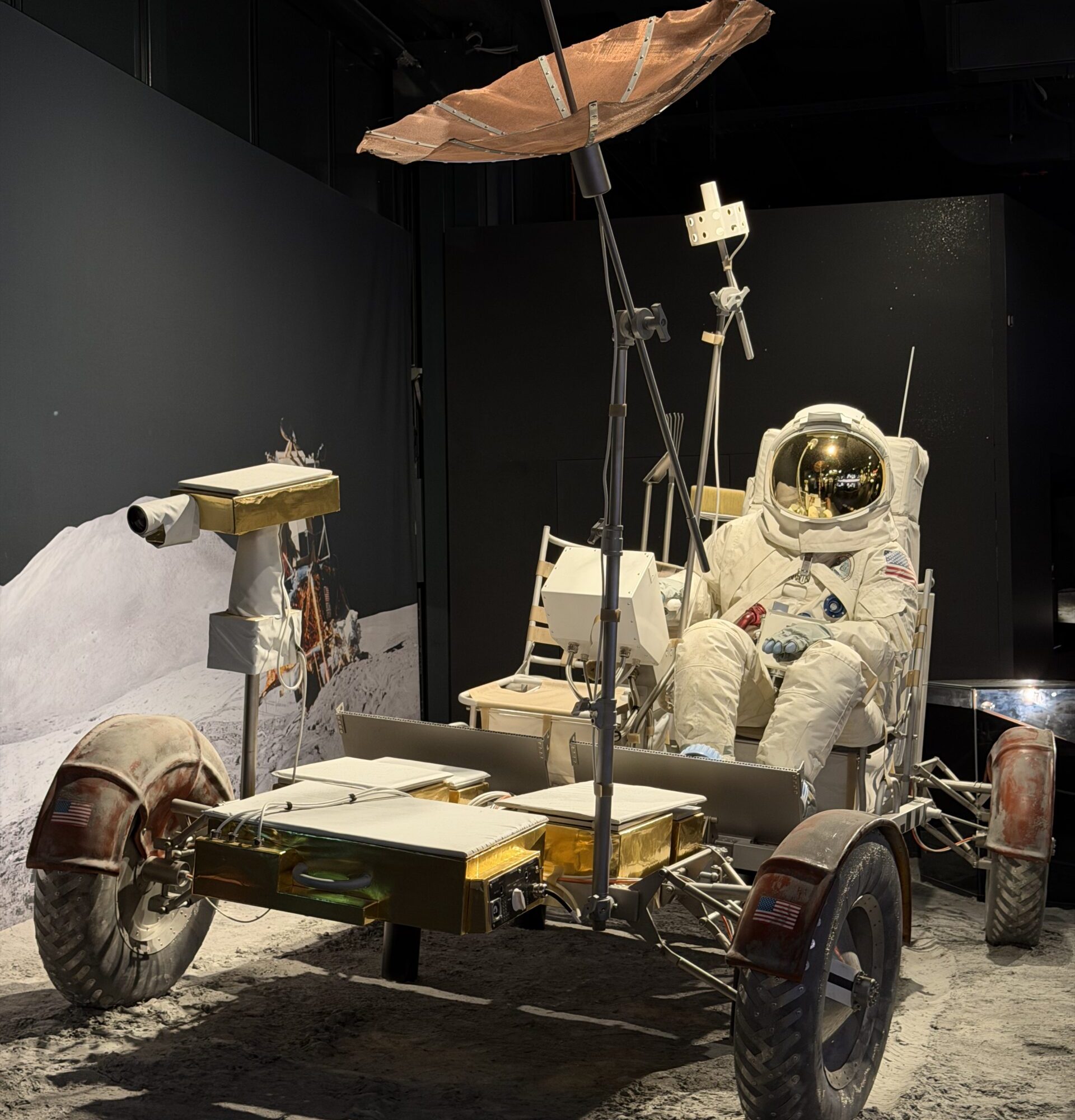 Lunar rover on a dusty exhibit stand with an astronaut in a white suit and gear nearby.