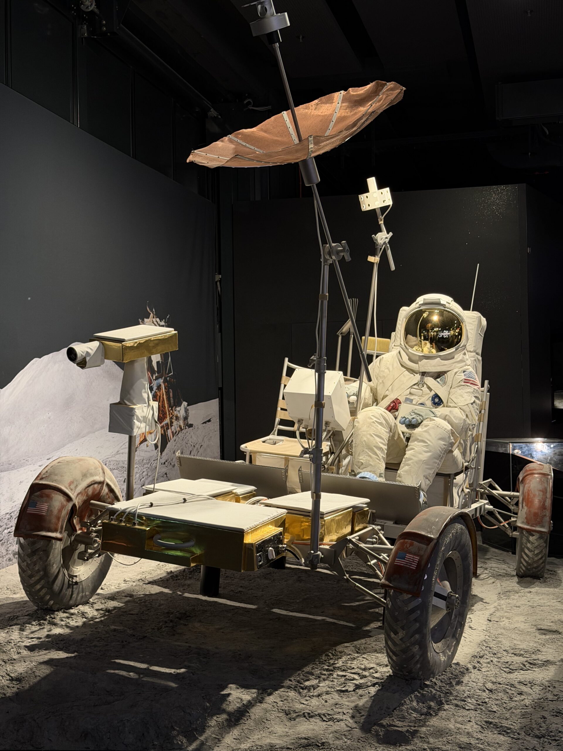Lunar rover on a dusty exhibit stand with an astronaut in a white suit and gear nearby.