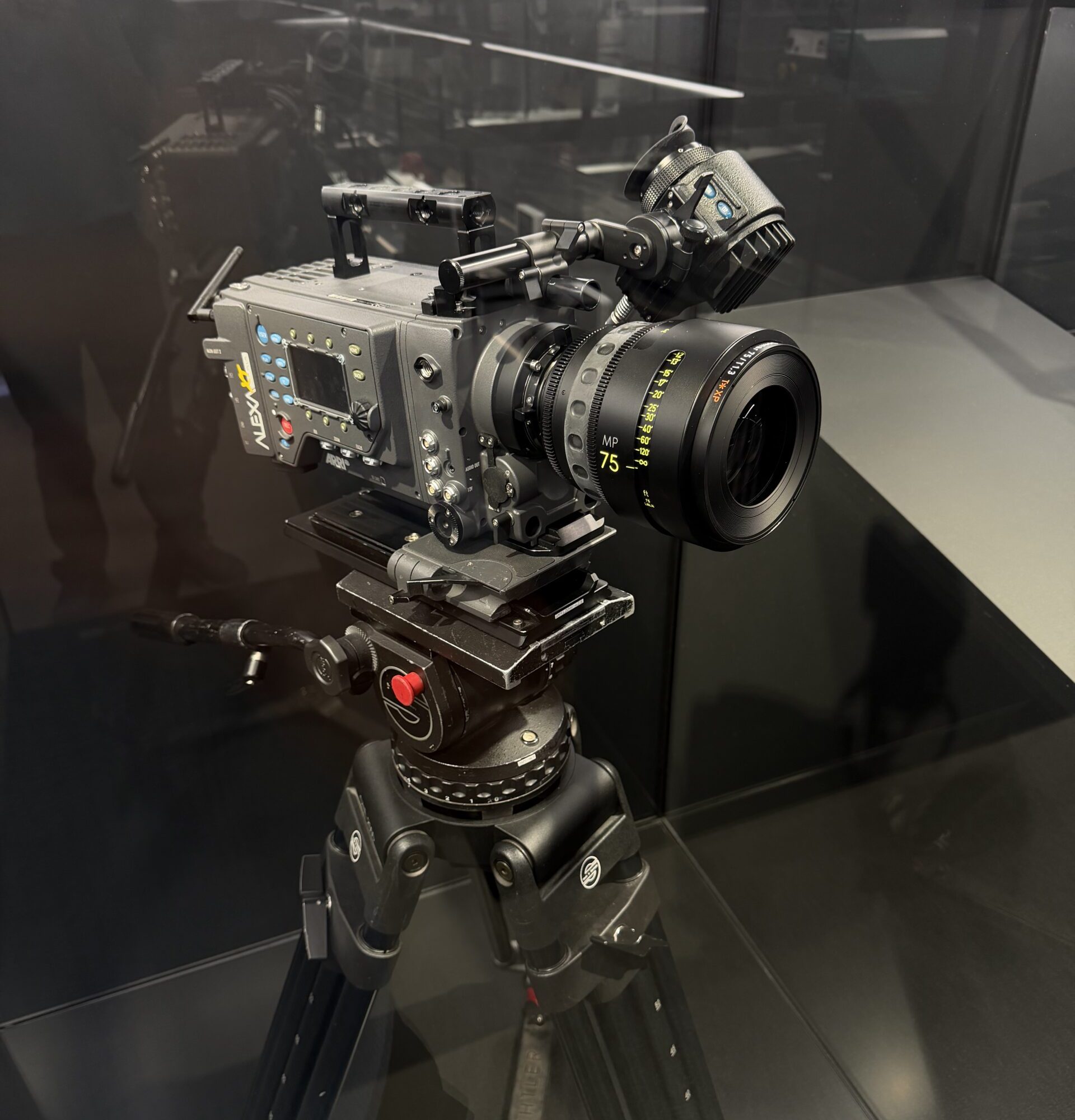 Professional cinema camera with a long lens mounted on a tripod inside a glass display case.