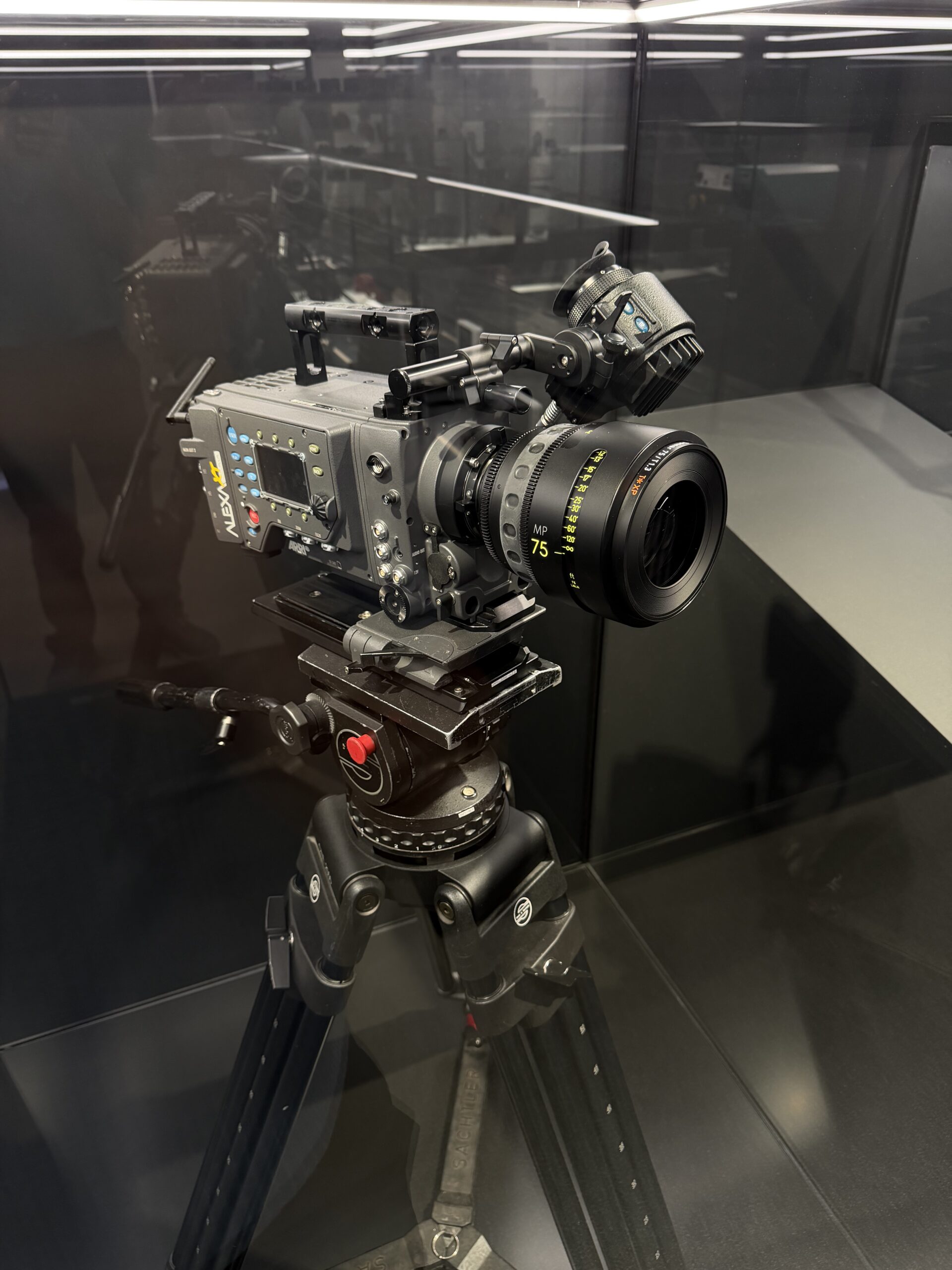 Professional cinema camera with a long lens mounted on a tripod inside a glass display case.