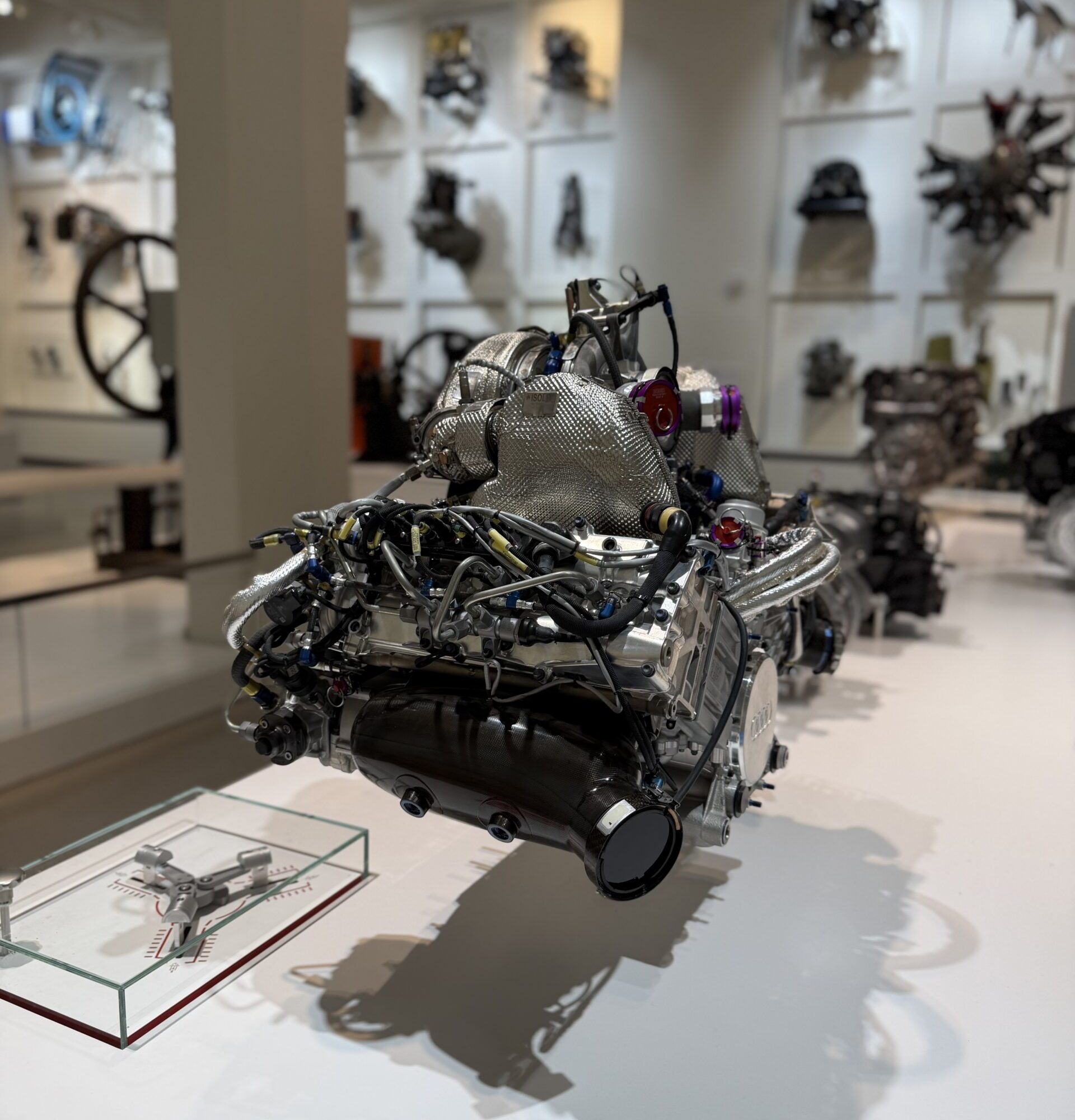 Close-up of a silver jet engine model with exposed wires on a display pedestal in a museum, with blurred engine mounts on white walls behind.