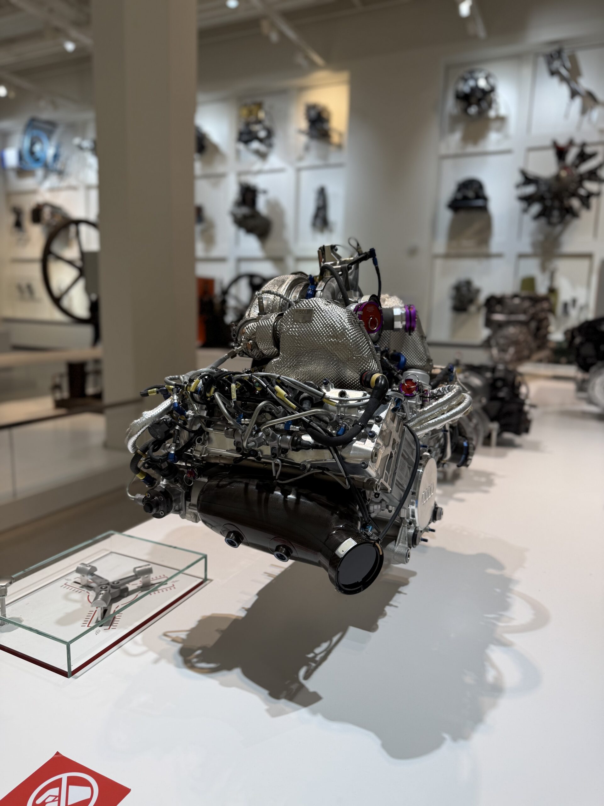 Close-up of a silver jet engine model with exposed wires on a display pedestal in a museum, with blurred engine mounts on white walls behind.