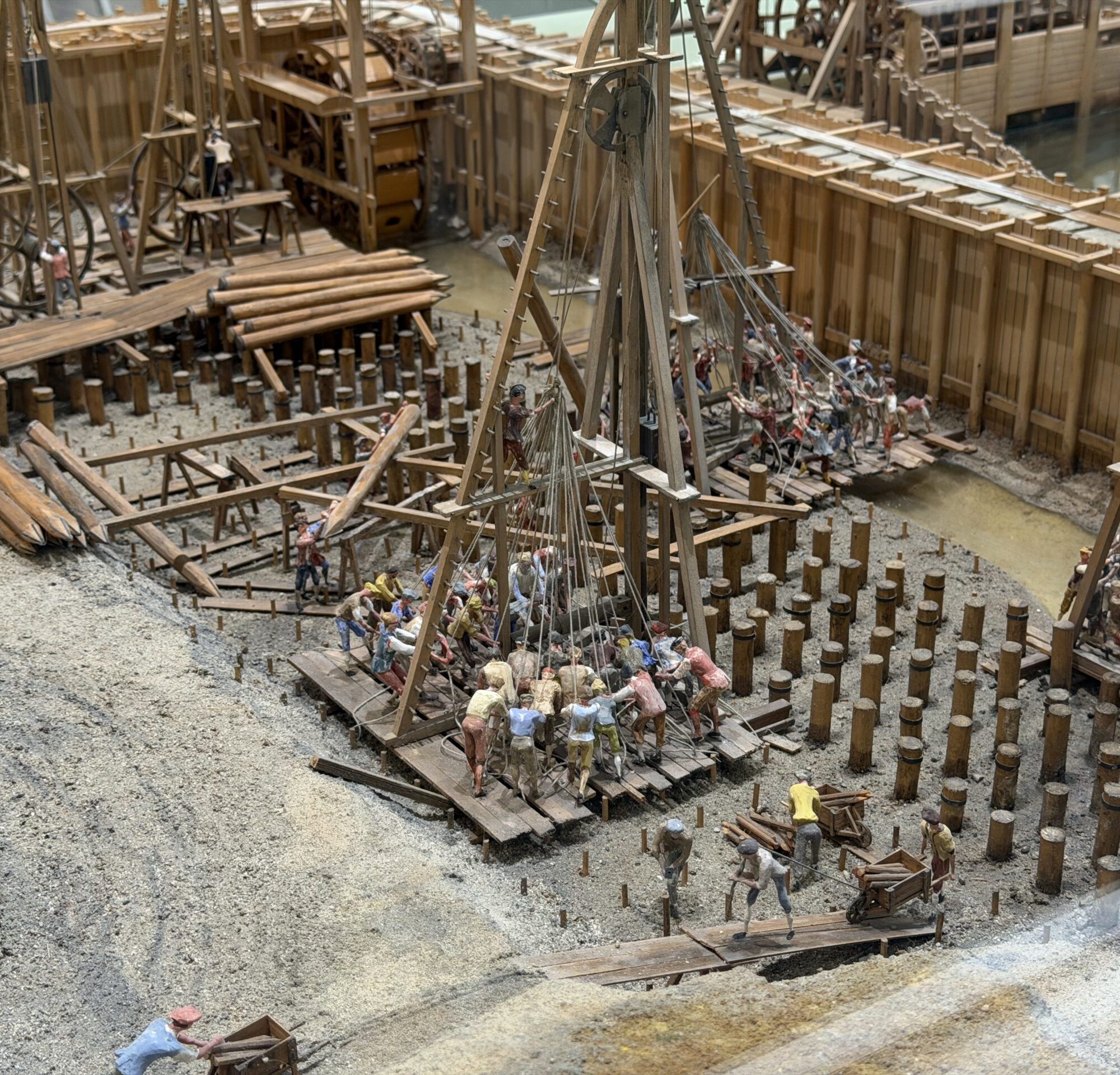 Scale-model construction site with wooden scaffolding, ramps, and dozens of worker figurines arranging logs and planks in a muddy excavation pit.