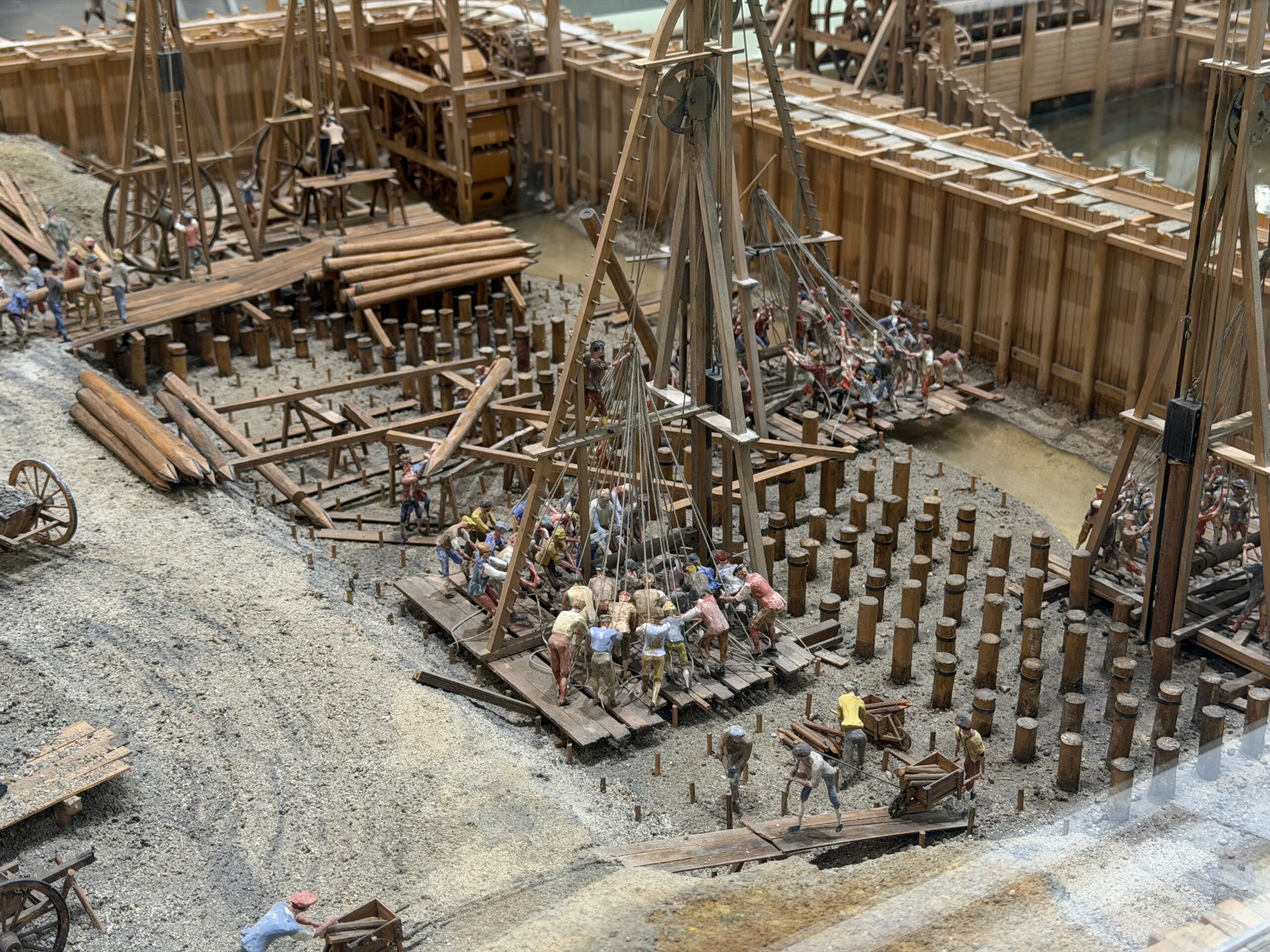 Scale-model construction site with wooden scaffolding, ramps, and dozens of worker figurines arranging logs and planks in a muddy excavation pit.