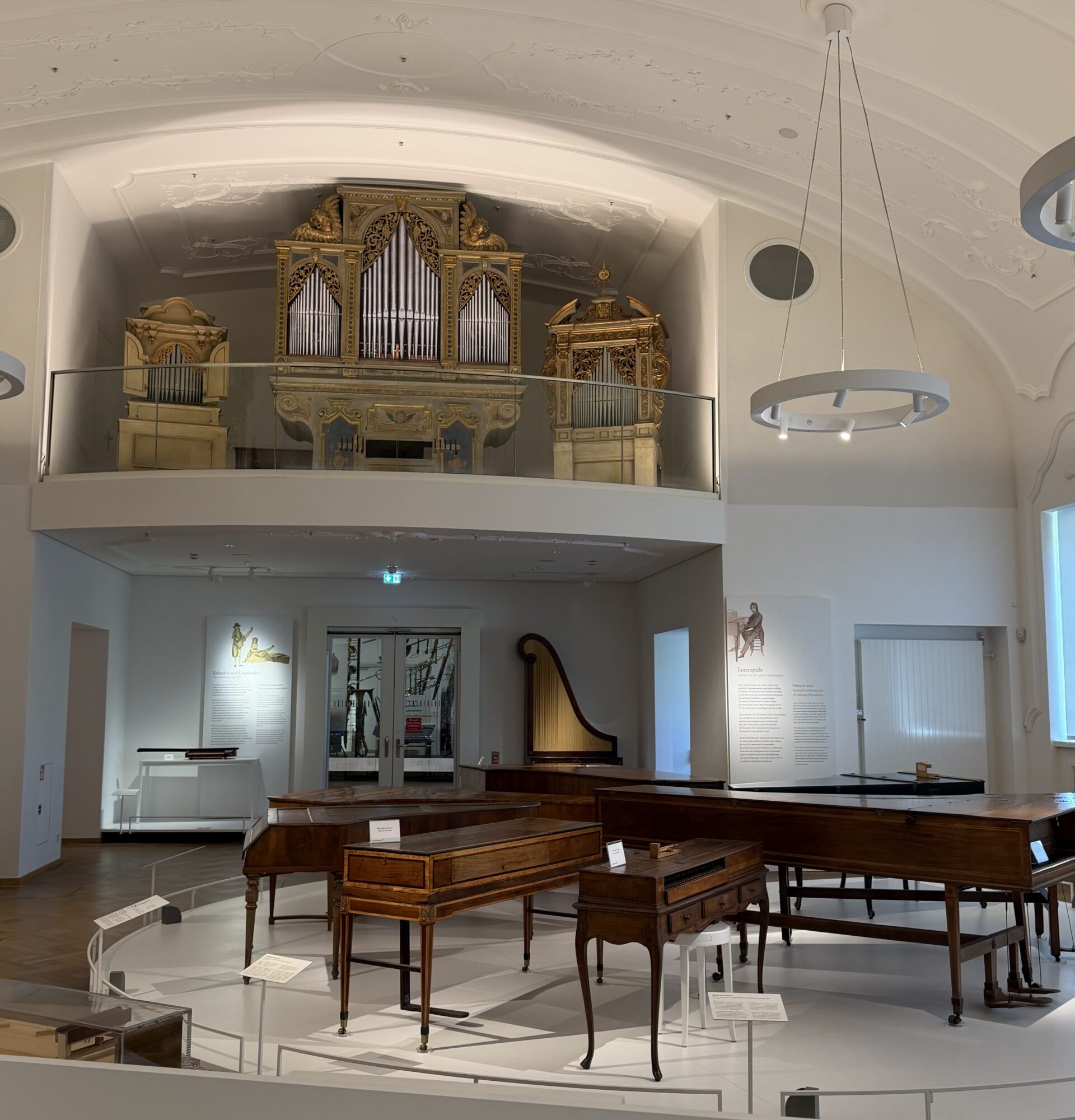 Museum gallery with an ornate, gold-leaf pipe organ on a mezzanine above wooden keyboard instruments nearby.