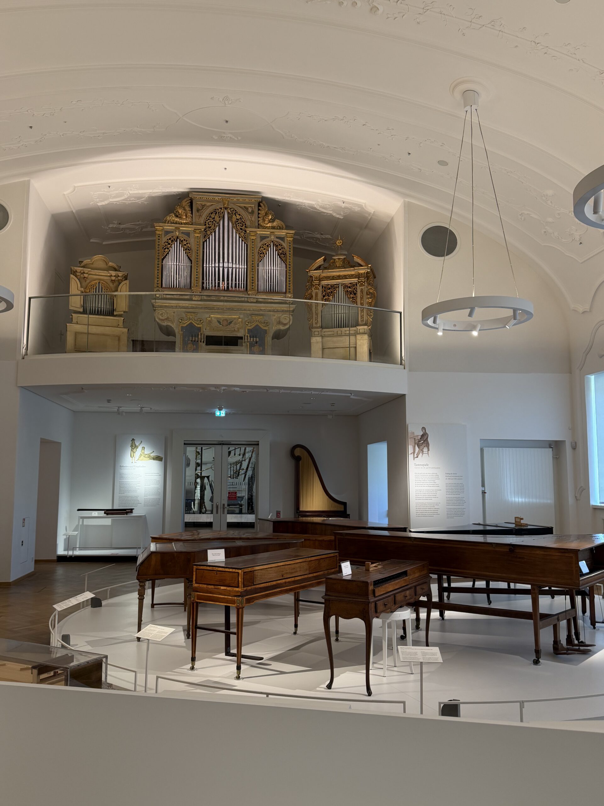 Museum gallery with an ornate, gold-leaf pipe organ on a mezzanine above wooden keyboard instruments nearby.