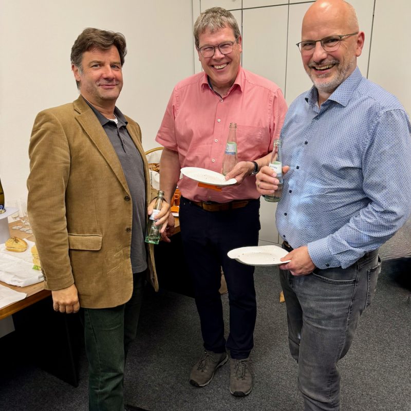 Three men stand together at a casual office gathering, each holding a bottle and a plate with snacks.
