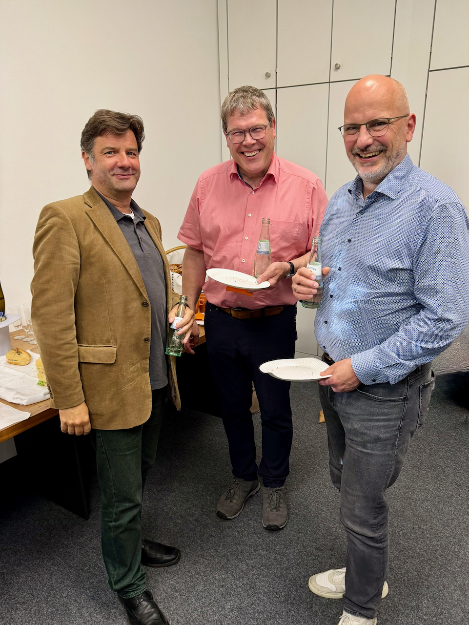 IMG_5110 Three men stand together at a casual office gathering, each holding a bottle and a plate with snacks.