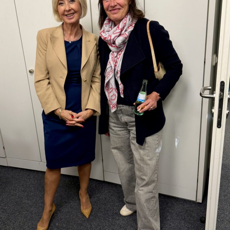 Two smiling women pose together indoors: left, a blonde in a beige blazer and navy dress; right, a brunette with a patterned scarf and dark coat, holding a bottle and a light-colored bag.