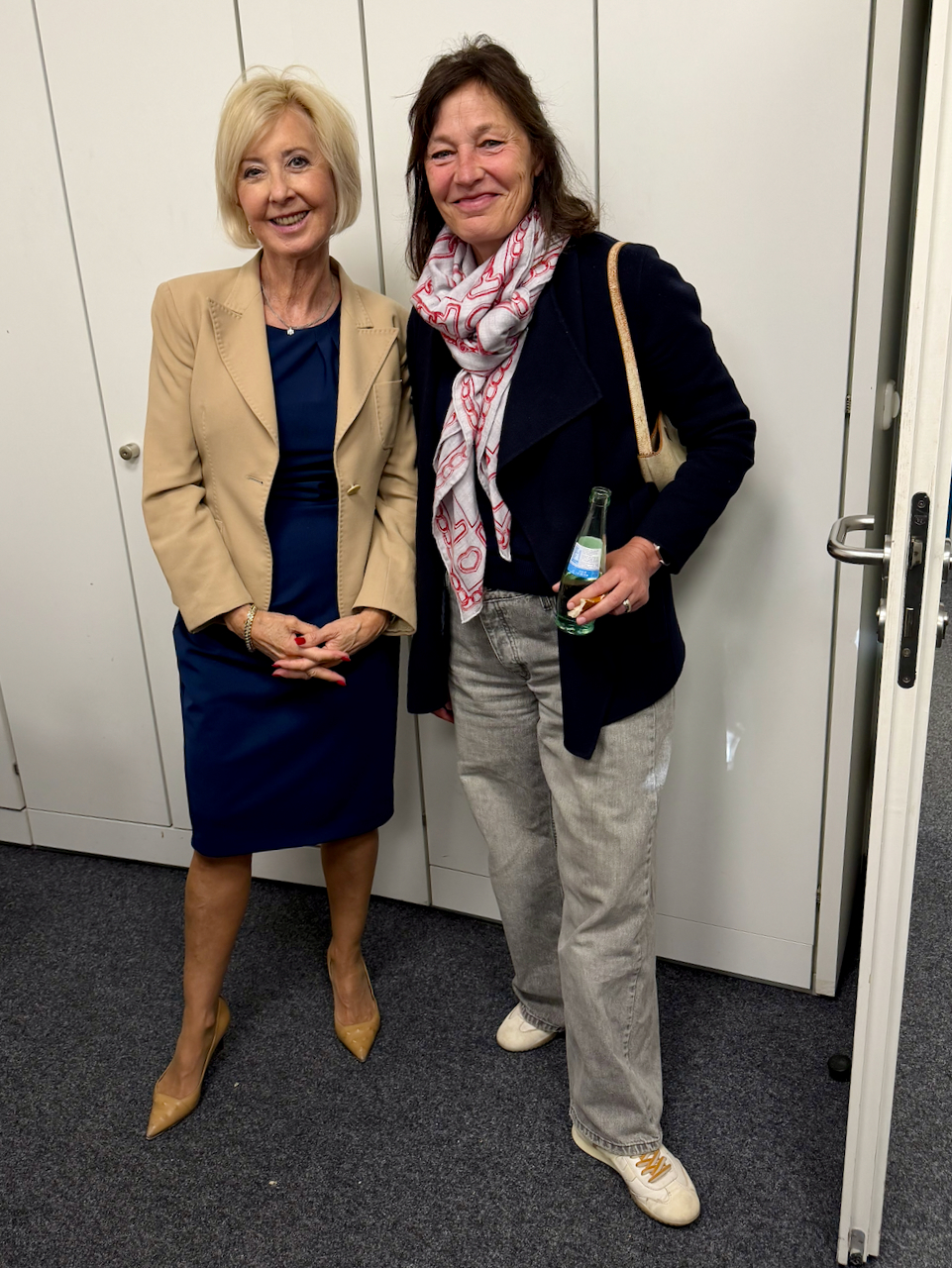 IMG_5113 Two smiling women pose together indoors: left, a blonde in a beige blazer and navy dress; right, a brunette with a patterned scarf and dark coat, holding a bottle and a light-colored bag.