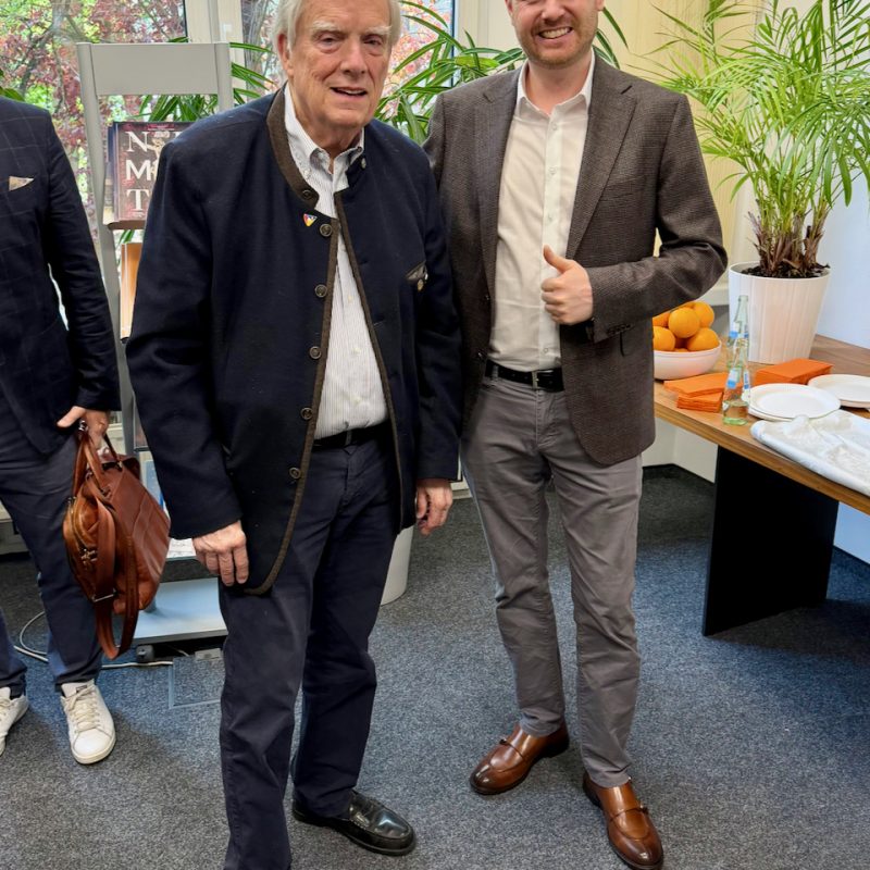 Two men pose for a photo at a bright indoor event with plants and a snack table in the background.