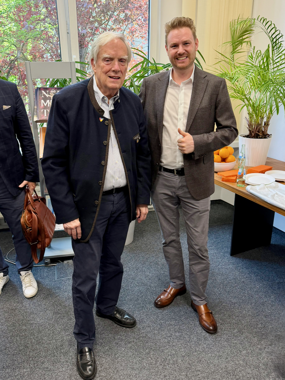 IMG_5115 Two men pose for a photo at a bright indoor event with plants and a snack table in the background.