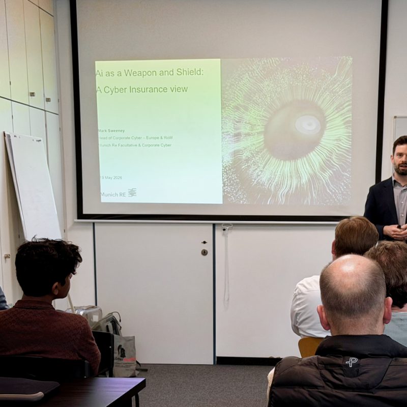 A man in a dark blazer giving a presentation in a classroom, with a projected slide about AI as a weapon and shield and a crowd listening.