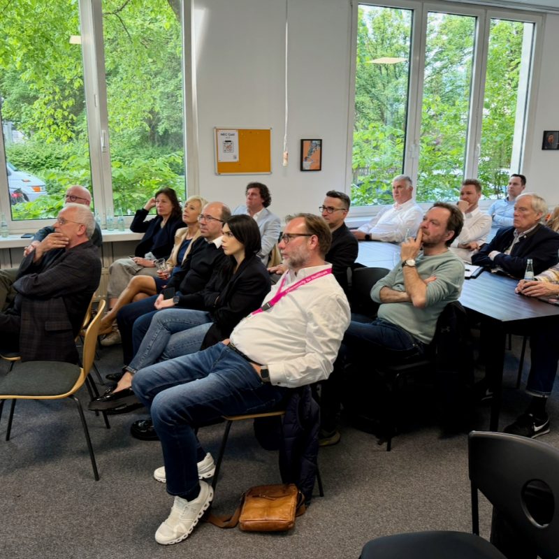 Audience of adults seated in a bright conference room watching a presentation with large windows and greenery outside.