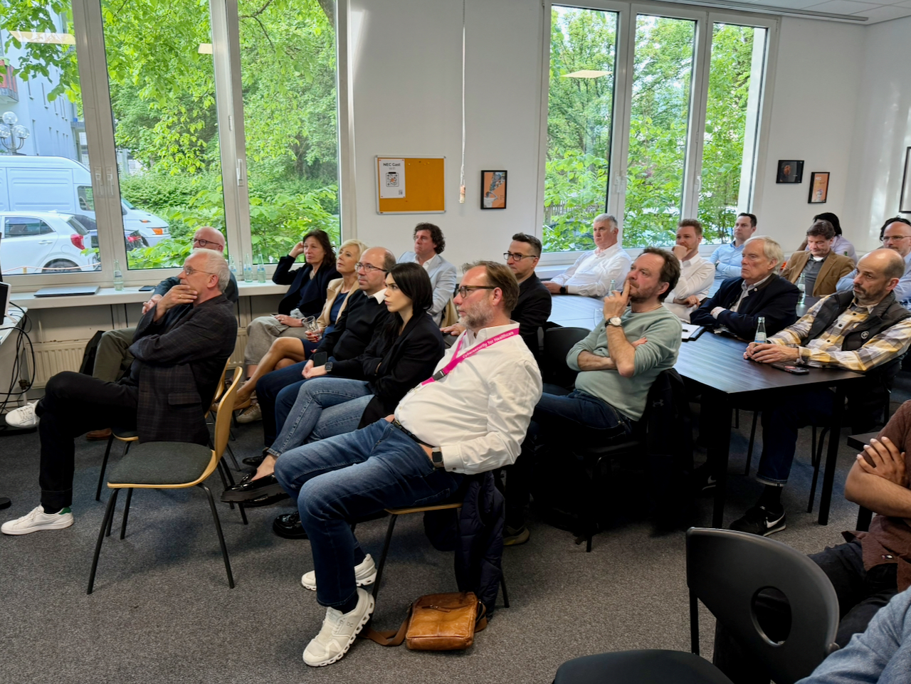 IMG_5126 Audience of adults seated in a bright conference room watching a presentation with large windows and greenery outside.