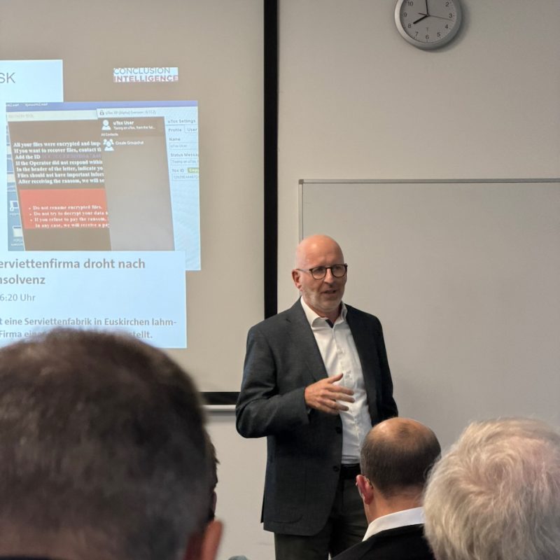 Bald man in a suit giving a presentation to an audience in a classroom, projector screen showing slides including the words 'CONCLUSION INTELLIGENCE' and German text, with a clock on the wall and a whiteboard to the side.