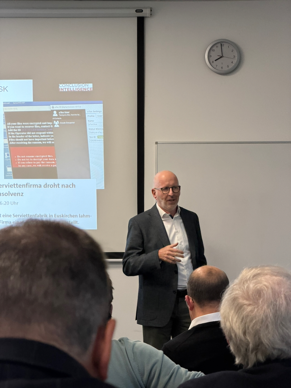 IMG_5133 Bald man in a suit giving a presentation to an audience in a classroom, projector screen showing slides including the words 'CONCLUSION INTELLIGENCE' and German text, with a clock on the wall and a whiteboard to the side.